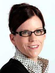 A white woman smiling with her brunette hair in a bun, wearing black rectangular glasses and black and white polka dot blouse.