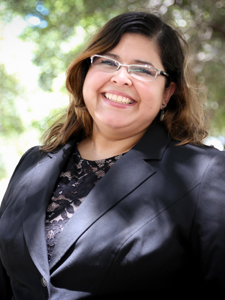 A Latina smiling with light brown hair, wearing silver squared glasses and a black blazer.
