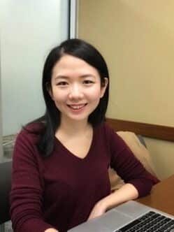 An Asian woman smiling with long, black hair, wearing a burgundy shirt and sitting at a wooden desk with a keyboard in front of her.