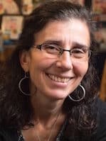 A white woman with dark brown, curly hair and frameless glasses smiling and wearing a black blouse and sweater.