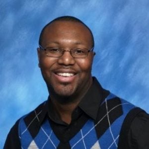 A Black man smiling with wire framed glasses, wearing a black dress shirt under a blue argyle vest with a blue background.