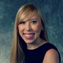 A white woman with long blonde hair and bangs swept to the side smiling, wearing a black sleeveless blouse.