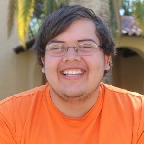 A Native man with short brown hair smiling, wearing frameless glasses and an orange t-shirt.