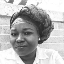 A Sudanese woman with a flat expression looking into the camera, wearing a head wrap and white blouse.