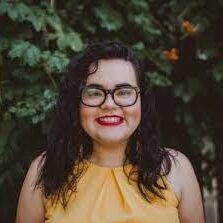 A Latina with shoulder length, curly black hair smiling with black-framed glasses and a yellow blouse.