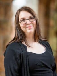 A white woman smiling with long brown hair and glasses, wearing a black blouse and cardigan.