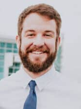 A white man with auburn brown, short hair and beard smiling, wearing a white dress shirt and blue tie.