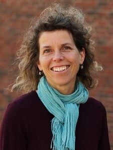 A white woman with curly, dark blonde hair smiling, wearing a teal scarf and purple shirt.