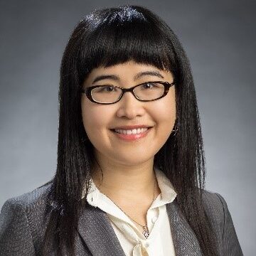An Asian woman smiling with shoulder-length, black hair and bangs, wearing black glasses, a white blouse, and grey blazer.