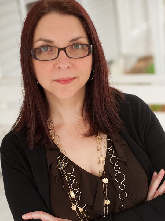 A white woman with long auburn hair and brown rectangular glasses looking at the camera, wearing a brown blouse with gold necklaces and black sweater.