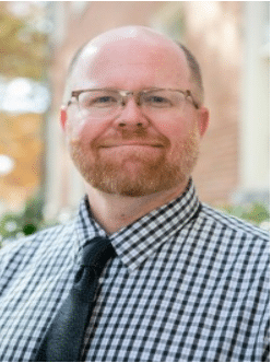 A bald, white man with an auburn beard and rectangular glasses smiling, wearing a black and white checked shirt and black tie.