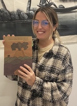 A Spanish woman with dark blonde hair and rounded glasses smiling in a plaid coat and holding up a book. 