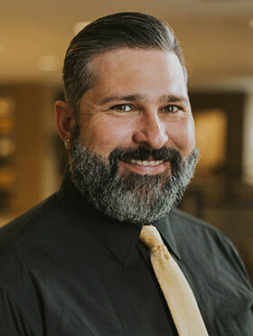 Chad Rose, a white man with brown, trimmed hair, with a dark brown and silver beard in a black button up shirt with a gold tie.