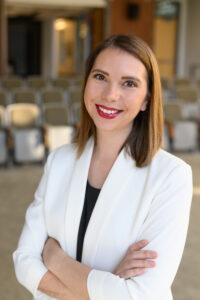 A white woman with shoulder-length light brown hair smiling and wearing a white blazer with a black shirt underneath. .