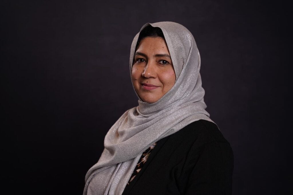 A professional headshot of Zohra Asad, a woman from Pakistan wearing a gray head wrap and a black sweater.
