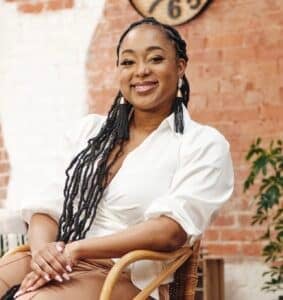 A professional image of Auriel Jasper-Morris, a black woman with braided hair in a white blouse and tan pants sitting down.